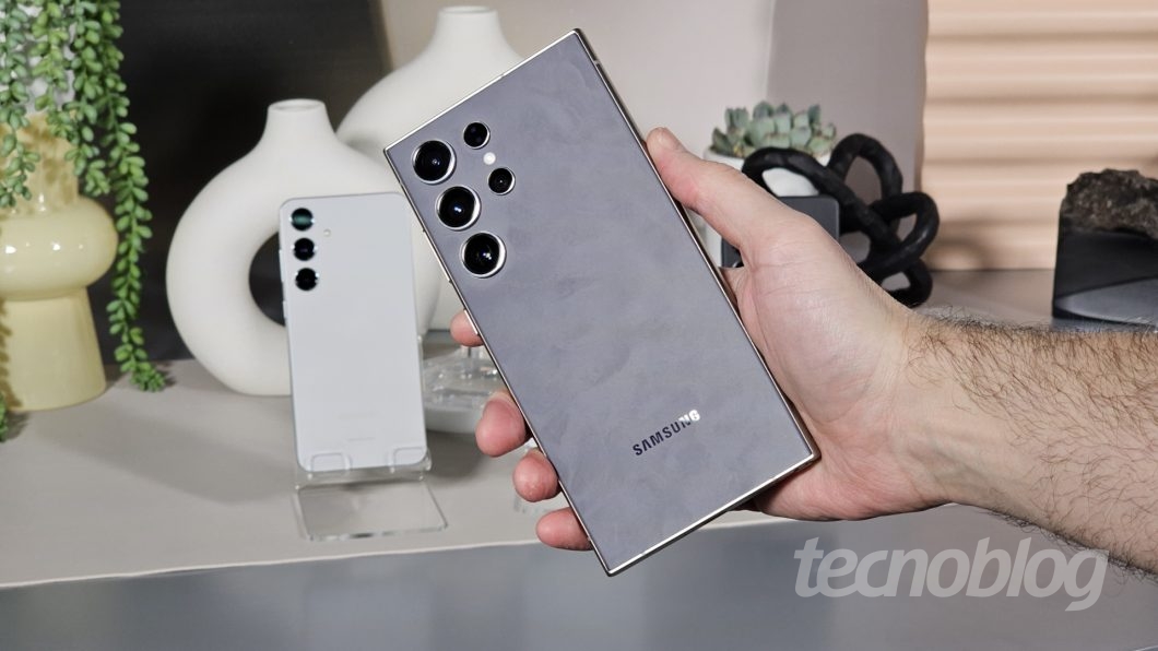 M&atilde;o segurando smartphone, ao fundo outro smartphone em um suporte de acr&iacute;lico sobre uma mesa