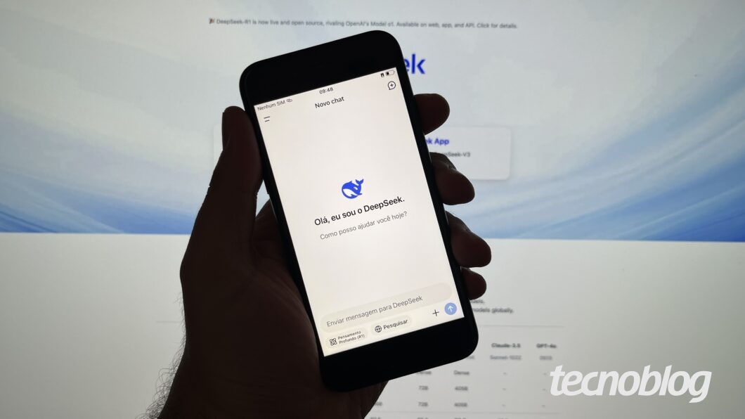 M&atilde;o segurando celular com o app do DeepSeek aberto. Ao fundo, o site do DeepSeek em um monitor.