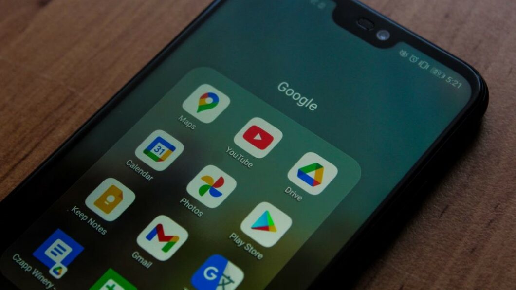 Smartphone sobre uma superf&iacute;cie de madeira, exibindo uma pasta nomeada &ldquo;Google&rdquo; na tela. Dentro da pasta, h&aacute; v&aacute;rios aplicativos do Google, incluindo Google Maps, YouTube, Google Drive, Google Calendar, Google Photos, Play Store, Keep Notes e Gmail.