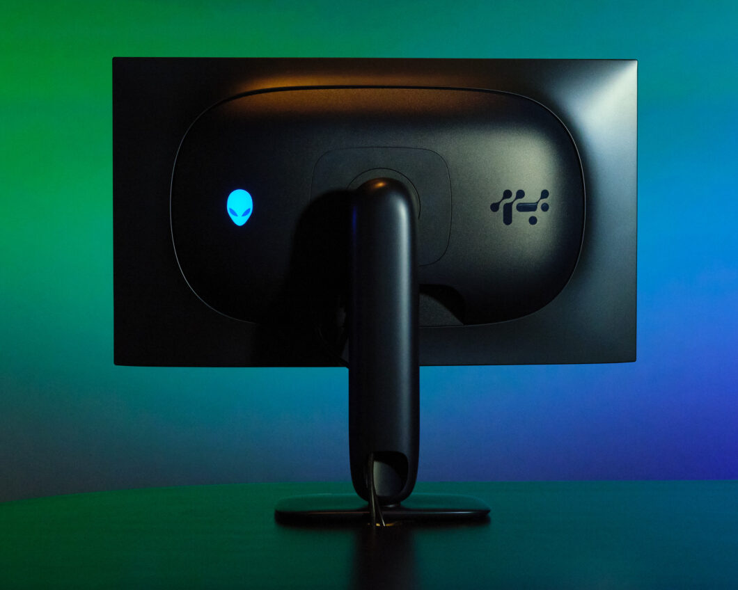Parte traseira de um monitor Alienware com acabamento preto fosco, logotipo iluminado em azul e um suporte central robusto. O fundo apresenta uma ilumina&ccedil;&atilde;o gradiente em tons de azul e verde.