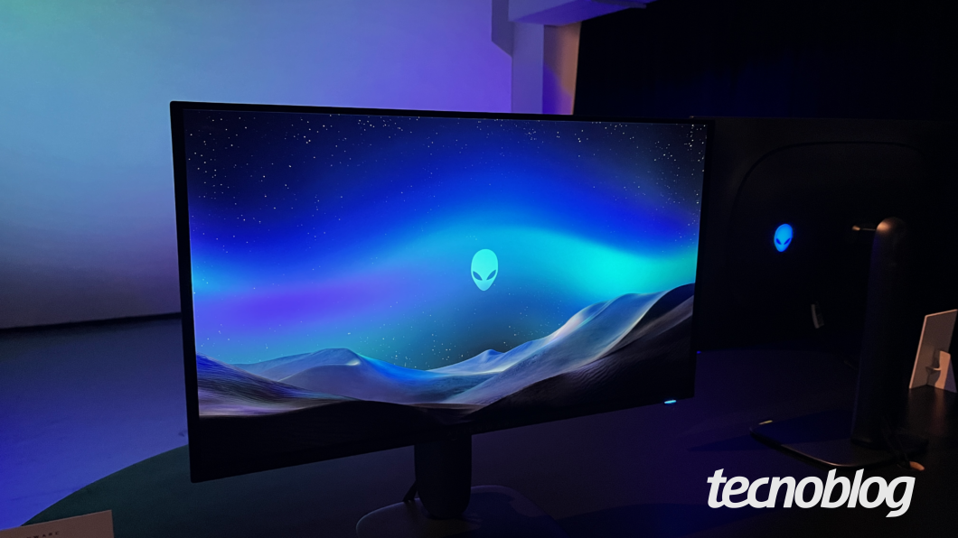 Monitor Alienware exibindo uma imagem com tema espacial, apresentando o logotipo da marca no centro da tela. O ambiente tem ilumina&ccedil;&atilde;o azulada e h&aacute; outro monitor semelhante ao fundo.
