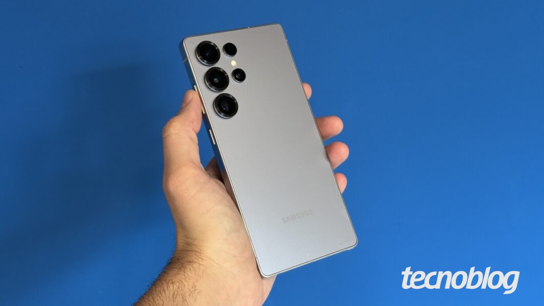 Uma m&atilde;o segura um smartphone Samsung de cor prateada contra um fundo azul. O aparelho possui um design premium com cantos retos e uma configura&ccedil;&atilde;o de c&acirc;mera traseira composta por quatro lentes dispostas verticalmente no canto superior esquerdo. O logo da Samsung est&aacute; gravado na parte inferior traseira do dispositivo.