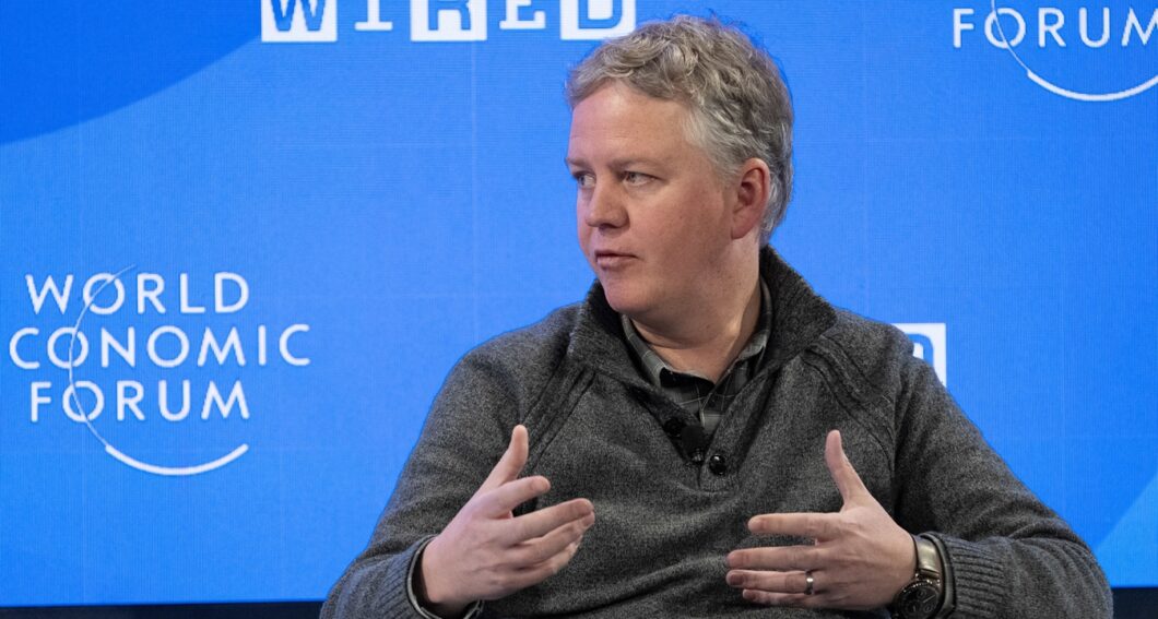Imagem colorida mostra Matthew Prince, CEO da Cloudflare. Ele &eacute; um homem de cabelos grisalhos curtos, est&aacute; posicionado no centro da imagem, falando e gesticulando com as m&atilde;os abertas. Ele veste um su&eacute;ter cinza escuro sobre uma camisa preta. O fundo &eacute; uma tela azul vibrante com as palavras "WORLD ECONOMIC FORUM" impressas em letras brancas grandes &agrave; esquerda.