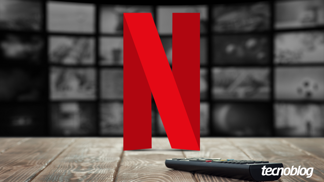 Imagem mostra o logo vermelho estilizado da Netflix, um "N" grande com uma curva na parte inferior. Abaixo e ligeiramente &agrave; direita do logo, um controle remoto preto est&aacute; parcialmente vis&iacute;vel. Ao fundo, uma parede composta por v&aacute;rias telas de televis&atilde;o desfocadas exibe diversas cenas de filmes e s&eacute;ries em tons de cinza. Na parte inferior direita, o logotipo do "Tecnoblog" &eacute; vis&iacute;vel.