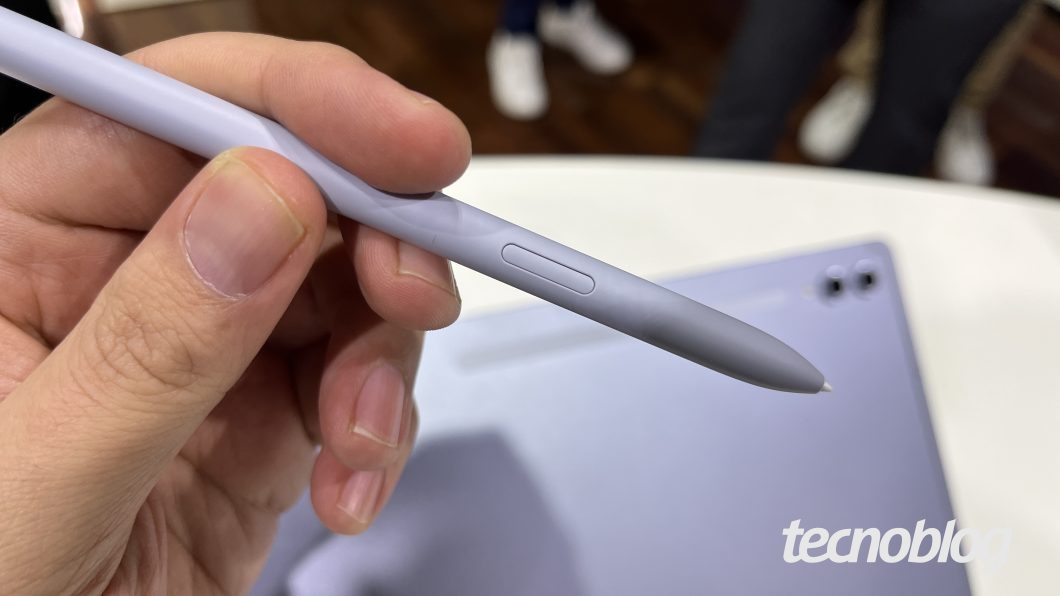 Mão segura uma caneta cinza. Ao fundo, um tablet.