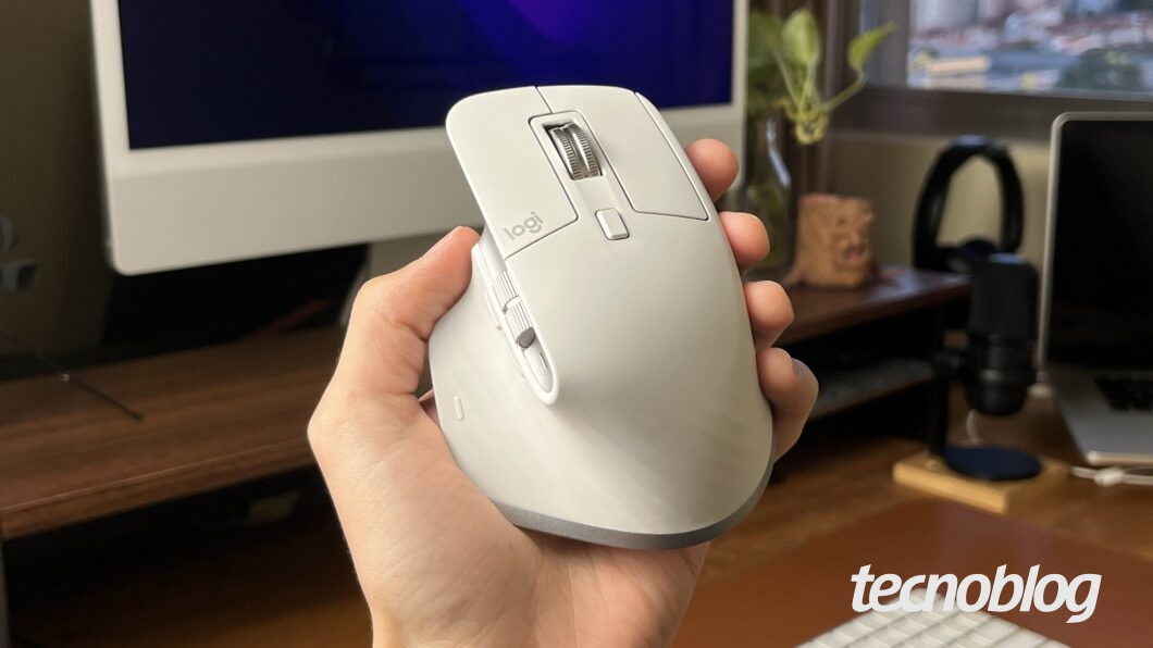 Uma mão segura um mouse ergonômico Logitech MX Master 3S branco. O mouse possui botões laterais e scroll. Ao fundo, um monitor e plantas em um ambiente de escritório. No canto inferior direito, o logo "tecnoblog".
