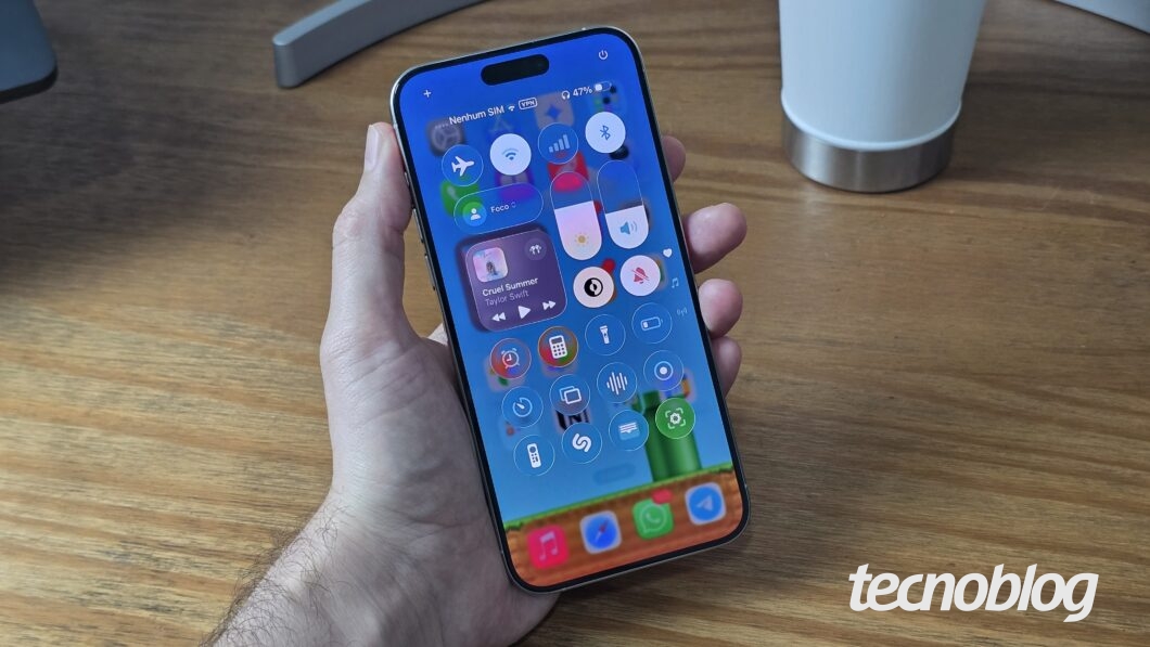 Imagem mostra uma mão segurando um iPhone com o sistema iOS 26 beta. A mão e o aparelho estão sobre uma mesa de madeira. Na parte inferior direita, o logotipo do "tecnoblog" é visível.