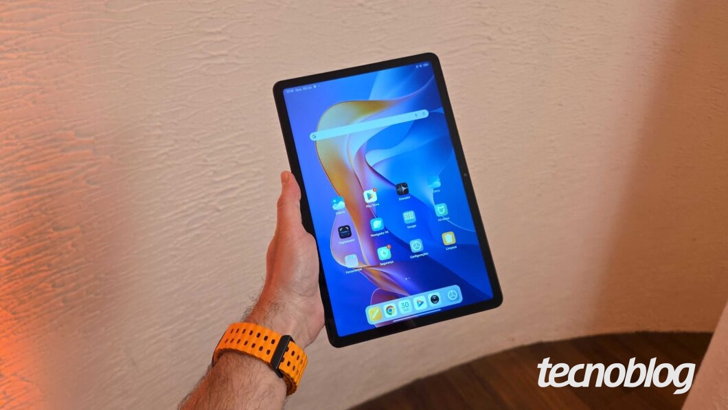 Uma mão segura um tablet Redmi Pad 2 de cor cinza escuro, com a tela ligada, exibindo os apps. O fundo é uma parede texturizada de cor clara e um piso de madeira. Na parte inferior direita, a marca d'água do "tecnoblog" é visível.