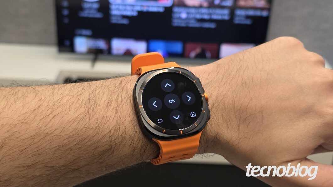 Braço com relógio smartwatch de pulseira laranja, tela exibe controles direcionais com botões "OK" e ícones de setas, casa e voltar.