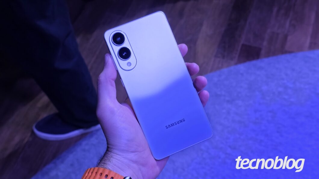 Uma m&atilde;o segura um smartphone Samsung Galaxy S25 Edge de cor prateada, com a parte traseira voltada para cima. O celular possui duas c&acirc;meras circulares pretas alinhadas verticalmente no canto superior esquerdo. O nome "SAMSUNG" &eacute; vis&iacute;vel na parte inferior da traseira. O fundo est&aacute; desfocado em tons de roxo e azul, com o p&eacute; de uma pessoa aparecendo &agrave; esquerda. Na parte inferior direita, o logotipo do "tecnoblog" &eacute; vis&iacute;vel.