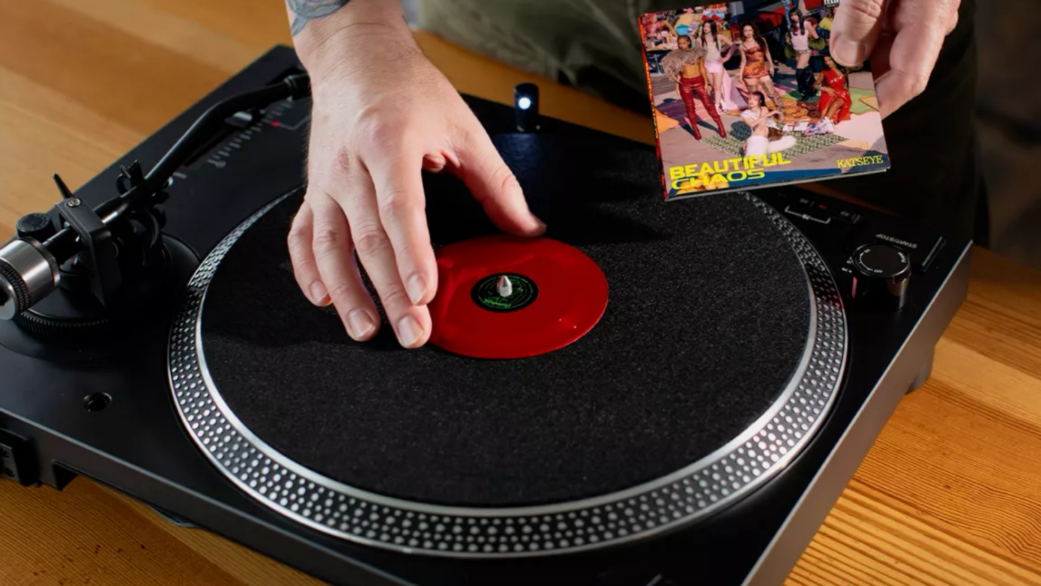 Mãos de uma pessoa com tatuagens no braço posicionando um pequeno disco de vinil vermelho no centro de um toca-discos preto. A outra mão segura a capa do disco, que é pequena e colorida. O toca-discos está sobre uma superfície de madeira.