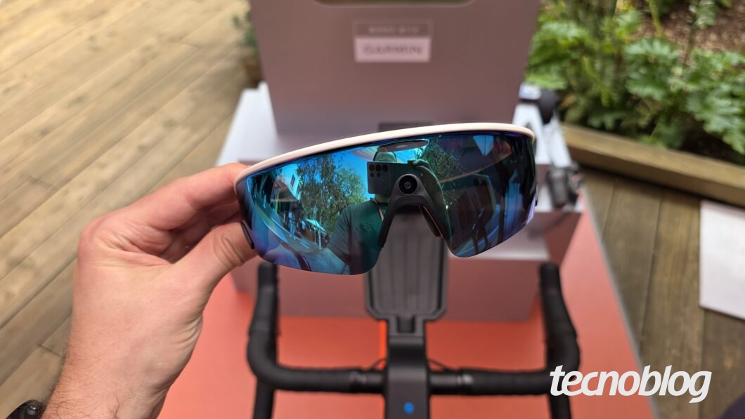 Imagem mostra um óculos smart Oakley Meta Vanguard segurado em uma mão. Na parte inferior direita, o logotipo do "tecnoblog".
