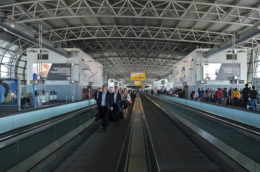 Vista interna de um terminal de aeroporto com teto alto em estrutura metálica e iluminação natural pelas laterais. No centro, há uma esteira rolante dupla para pedestres, com pessoas caminhando em ambas as direções, algumas com malas de mão. À esquerda e à direita, filas de passageiros e áreas de embarque. Painéis publicitários grandes com imagens e textos estão fixados nas paredes superiores. Placa amarela no fundo exibe "Vueling – FLY TO SPAIN".