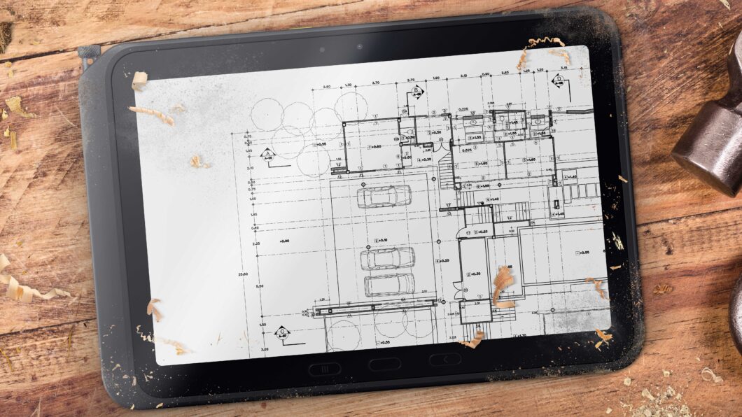 Tablet sobre mesa de madeira exibindo planta arquitetônica digital, cercado por lascas de madeira e uma ferramenta metálica, sugerindo um ambiente de marcenaria ou construção.