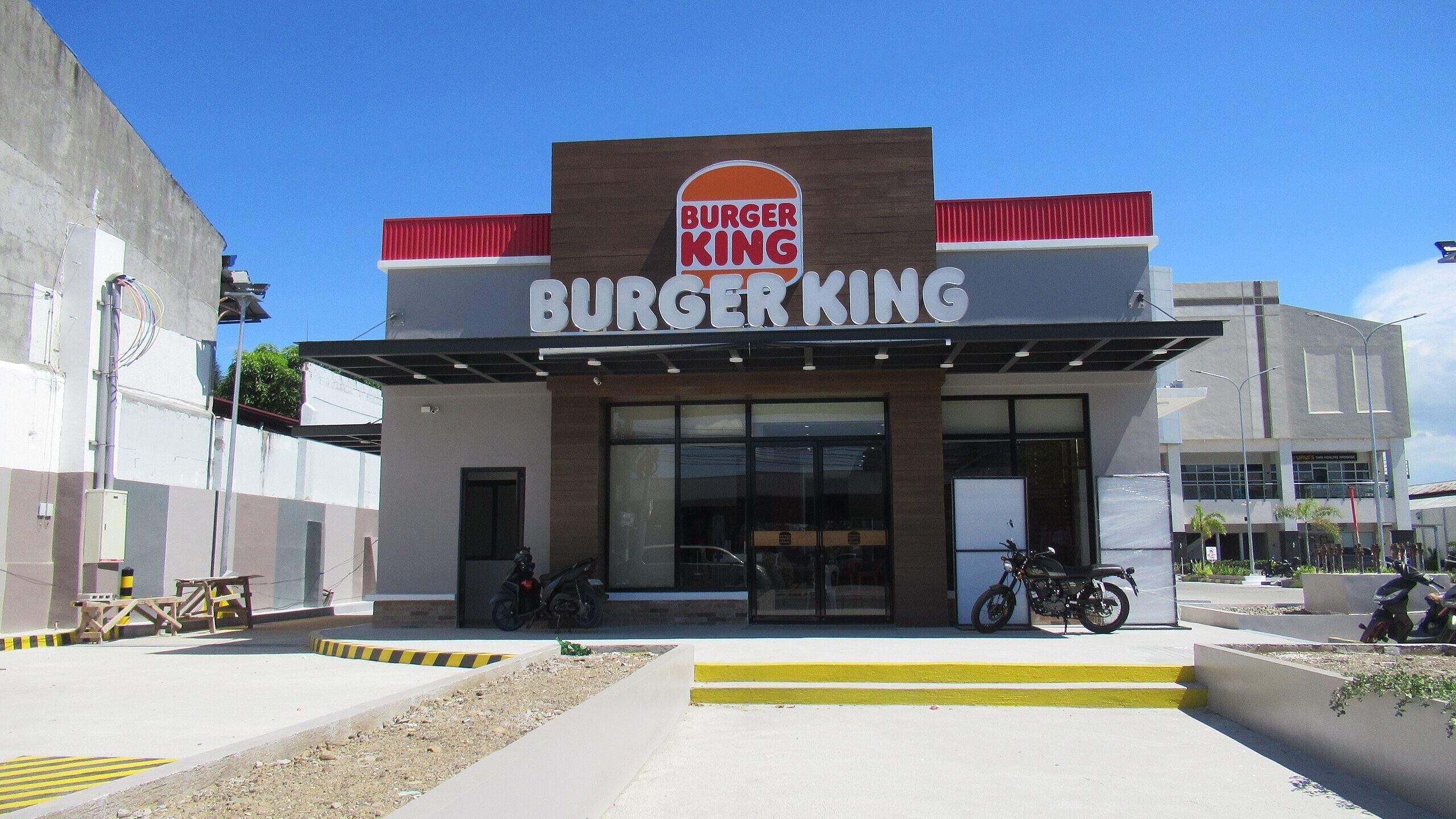 Fachada de uma unidade da rede Burger King em dia ensolarado. O pr&eacute;dio tem acabamento em tons de branco, cinza e marrom, com a logomarca "BURGER KING" em letras grandes brancas no centro da estrutura, e o logotipo circular com as palavras "BURGER KING" em vermelho sobre fundo laranja acima. Em frente &agrave; entrada de vidro, h&aacute; duas motocicletas estacionadas. &Agrave; esquerda, h&aacute; mesas de madeira e uma &aacute;rea de estacionamento com faixas amarelas e pretas.