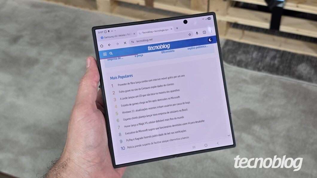M&atilde;o segurando smartphone dobr&aacute;vel aberto, exibindo a p&aacute;gina do Tecnoblog