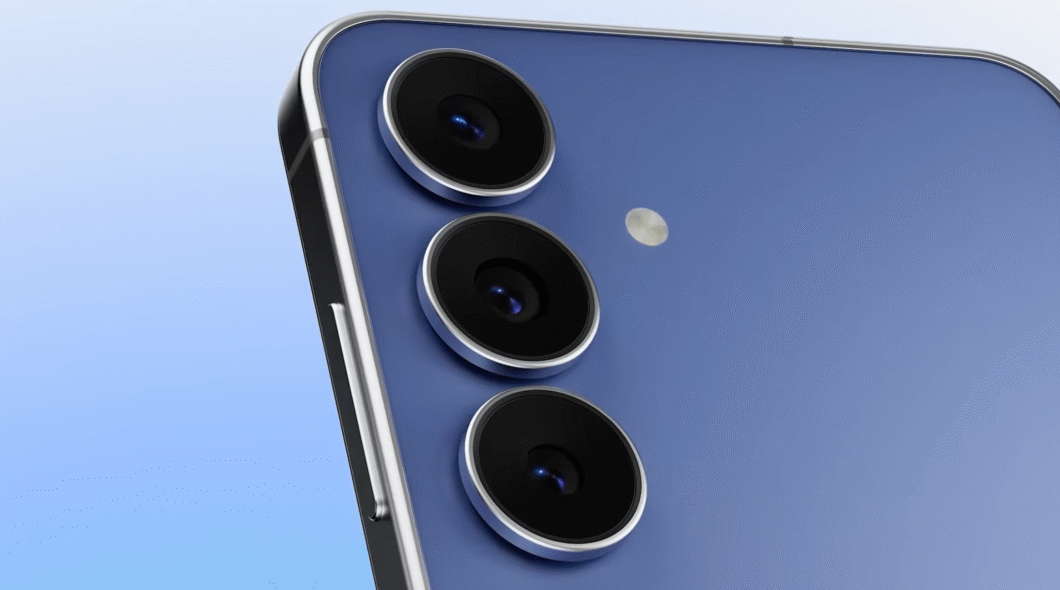 Uma imagem em close do smartphone Galaxy S25 FE azul-escuro. O aparelho tem um módulo de câmera vertical com três lentes alinhadas, cada uma cercada por um anel prateado. Ao lado das lentes, há um pequeno flash de LED. O fundo é um gradiente em tons de azul.