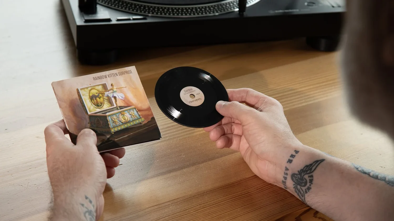 Imagem mostra uma mão segurando um Tiny Vinyl, disco de 4 polegadas, na mão direita, e a caixa do disco na mão esquerda. Os braços estão apoiados sobre uma mesa de madeira