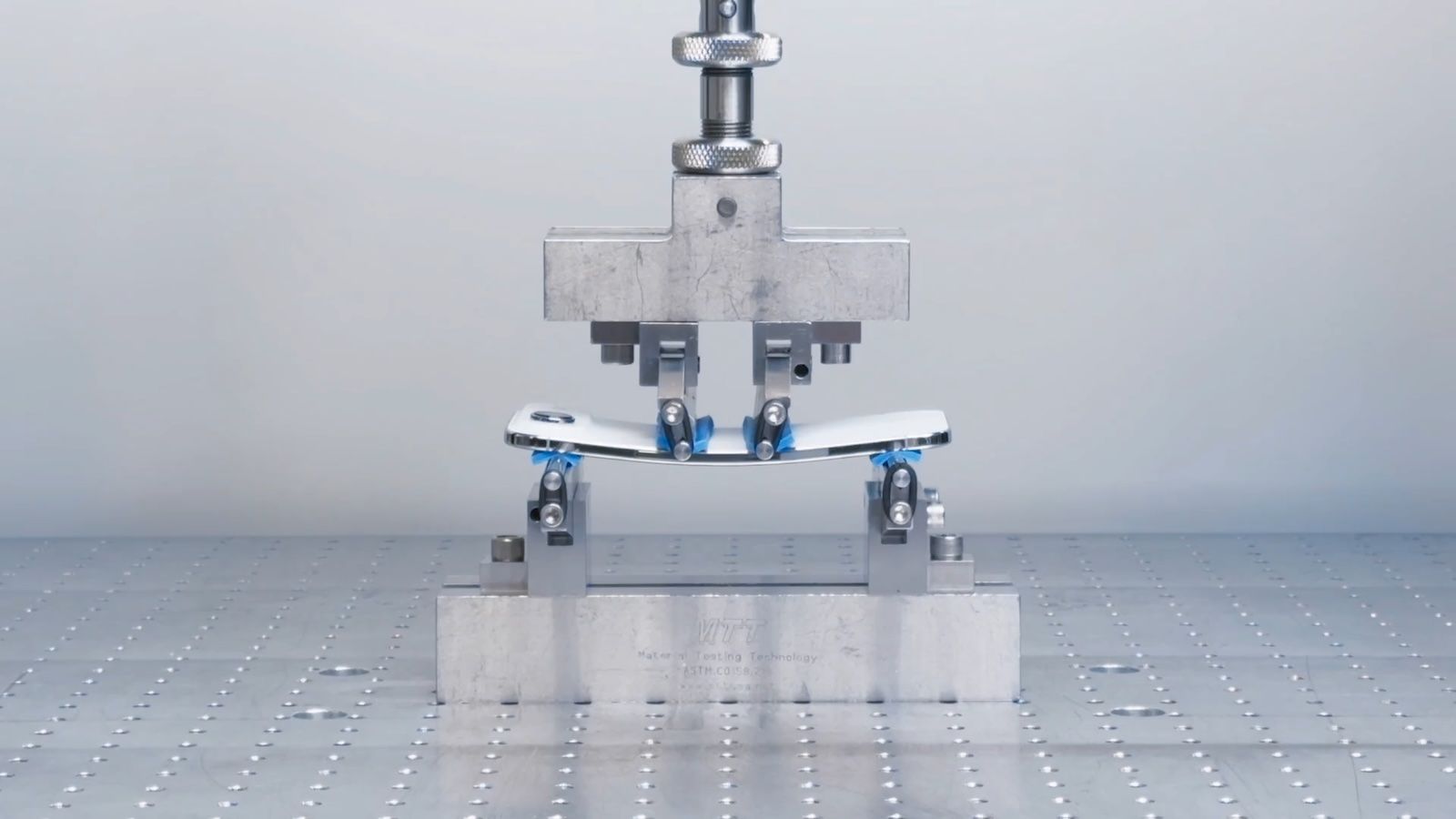 Uma fotografia de um teste de resistência mostra um smartphone iPhone Air sendo submetido a uma máquina de flexão. O aparelho está curvado para baixo no centro, indicando que está sendo dobrado. Ele é sustentado por duas bases metálicas e, no topo, uma haste de metal está pressionando o centro. As garras que seguram o aparelho têm detalhes azuis. O equipamento está sobre uma bancada metálica e a legenda do fabricante é visível.