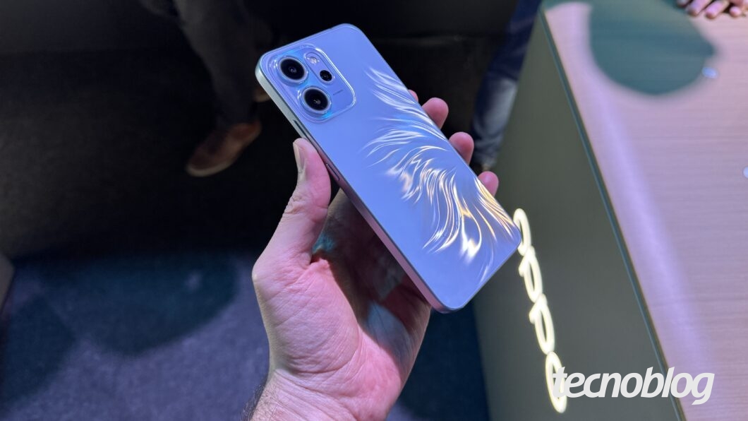 Foto de uma pessoa segurando um celular de cor azul-clara. O aparelho tem um acabamento texturizado na traseira que simula traços ou penas prateadas brilhantes. Na parte superior, é visível o módulo de câmera com duas lentes escuras. O celular é segurado sobre um balcão com o logotipo luminoso "Oppo". Na parte inferior direita, a marca d'água do "tecnoblog" é visível.
