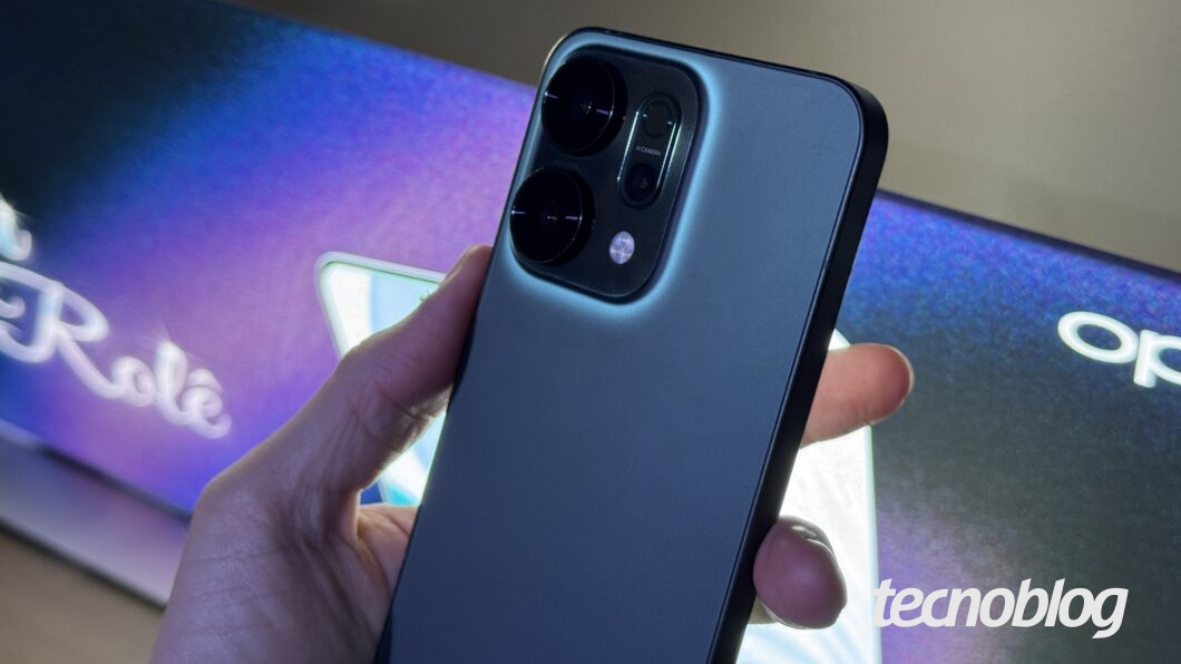 Foto em close-up da traseira de um celular Oppo Reno 14 de cor escura e fosco, segurado por uma mão. O celular possui um módulo de câmera grande com duas lentes circulares proeminentes e escuras, e o flash posicionado ao lado. A borda do módulo de câmera tem um brilho azulado que realça o aparelho. Ao fundo, é possível ver o logo luminoso "OPPO". Na parte inferior direita, a marca d'água do "tecnoblog".