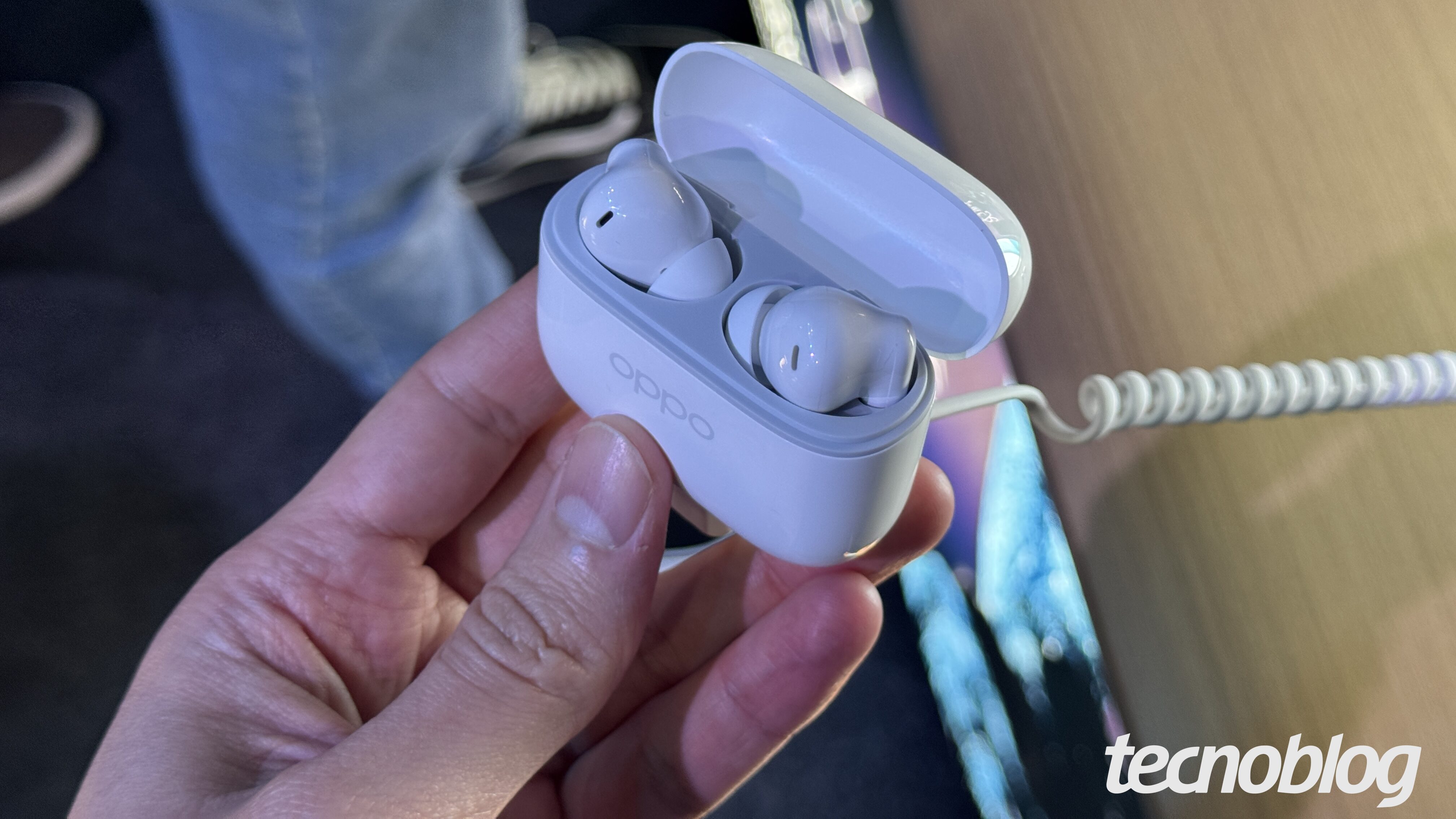 Foto em close-up de uma mão segurando um estojo branco aberto de fones de ouvido sem fio Oppo Enco Buds 3 Pro. O estojo tem o logo "oppo" em baixo relevo na frente e contém dois fones de ouvido brancos, no estilo intra-auricular. Na parte inferior direita, a marca d'água do "tecnoblog" é visível