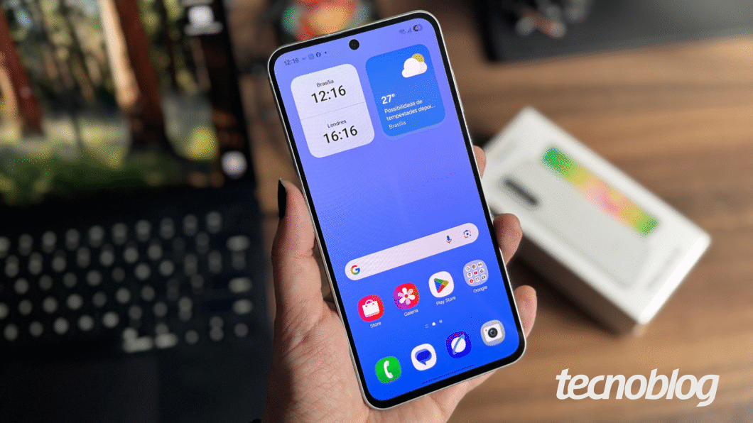 Imagem mostra mão segurando celular Samsung Galaxy A56 em primeiro plano, com fundo desfocado e caixa do aparelho ao fundo