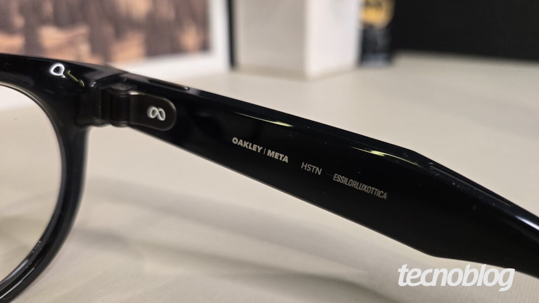 Foto em close-up de uma das hastes de óculos inteligentes de cor preta, focando nos elementos escritos em branco na parte interna. O texto visível é "OAKLEY | META", "HSTN" e "ESSILORLUXOTTICA".