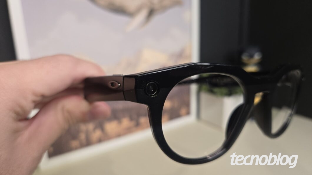 Foto de uma mão segurando, por uma das hastes, um par de óculos inteligentes Oakley Meta HSTN de armação preta e arredondada. No canto inferior direito da imagem, há o logotipo da marca "tecnoblog".
