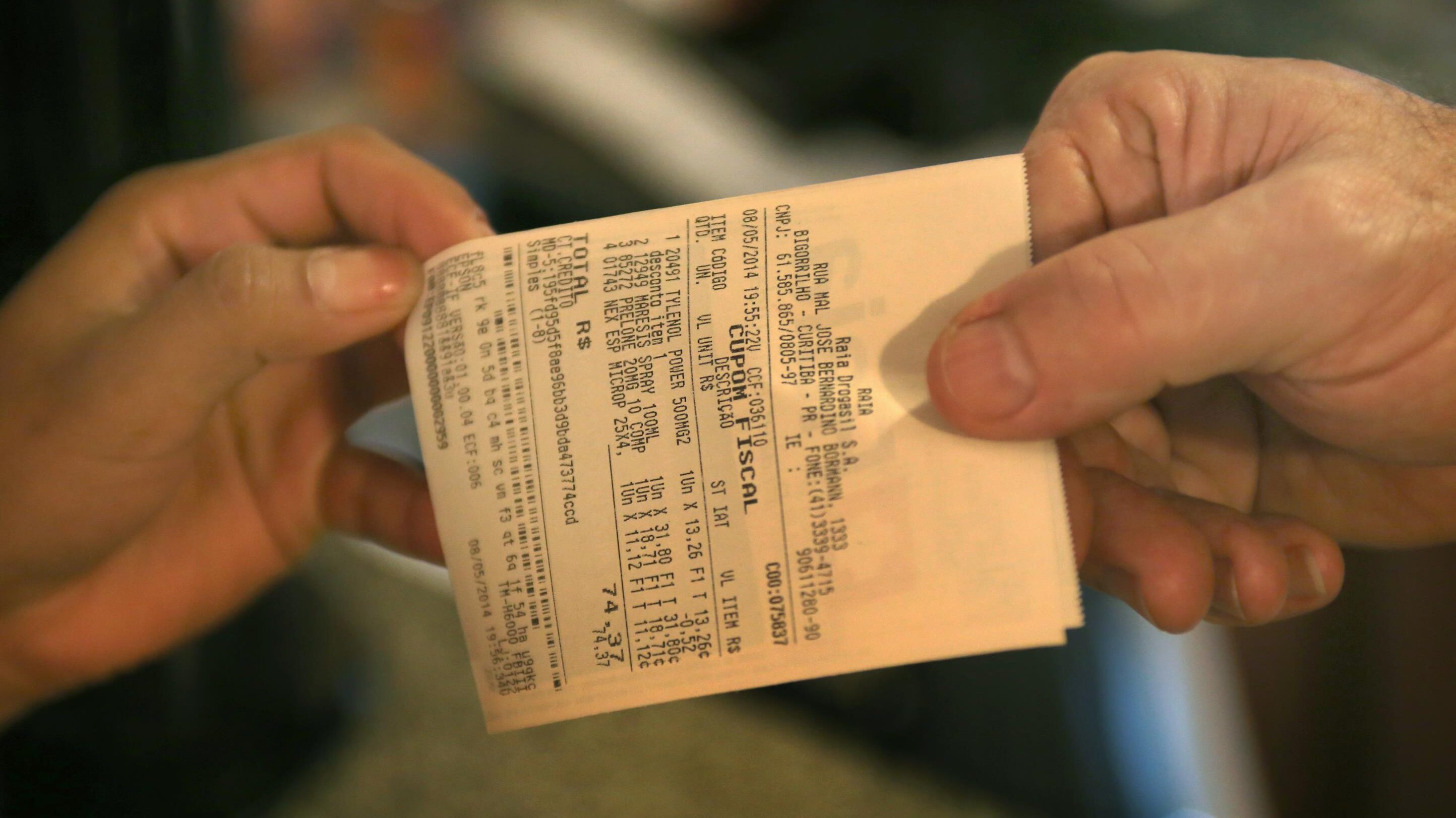 Foto em close-up de duas mãos segurando as pontas de um cupom fiscal ou nota de compra.