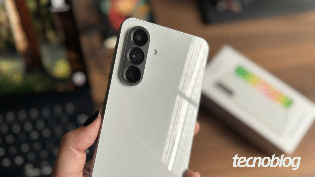 Imagem mostra mão segurando Samsung Galaxy A56 na cor, com a parte de traz à mostra. Traseira cinza exibe módulo preto com câmera tripla.