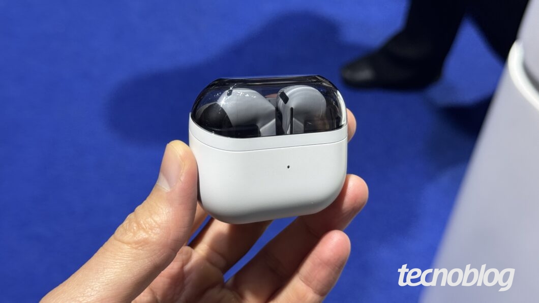 Uma mão segura um estojo de fones de ouvido sem fio. O estojo é branco com a tampa transparente e os dois fones são cinza-escuro. O logotipo "tecnoblog" está no canto inferior direito, em branco.