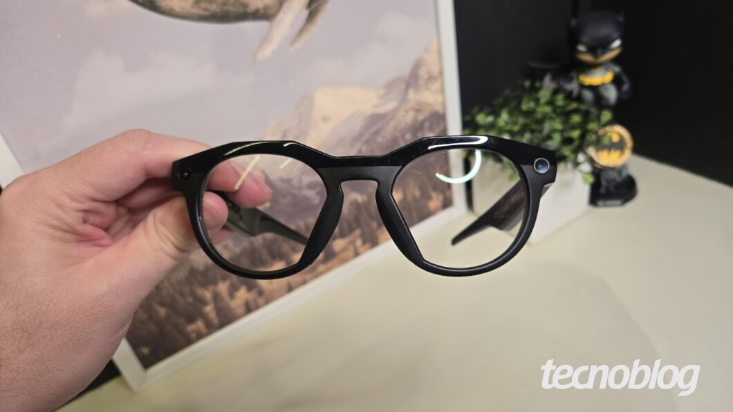 Foto de uma mão segurando, por uma das hastes, um par de óculos inteligentes Oakley Meta HSTN de armação preta e arredondada. No canto inferior direito da imagem, há o logotipo da marca "tecnoblog".