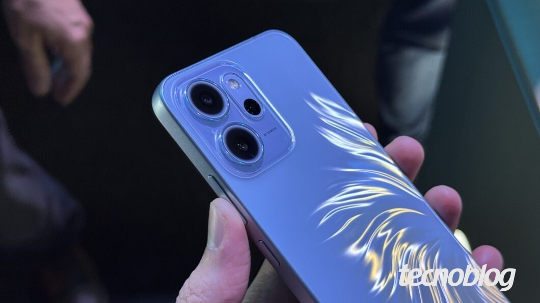 Foto de uma pessoa segurando um celular da Oppo de cor azul-clara. O aparelho tem um acabamento texturizado na traseira que simula traços ou penas prateadas brilhantes. Na parte superior, é visível o módulo de câmera com duas lentes escuras. Na parte inferior direita, a marca d'água do "tecnoblog" é visível.