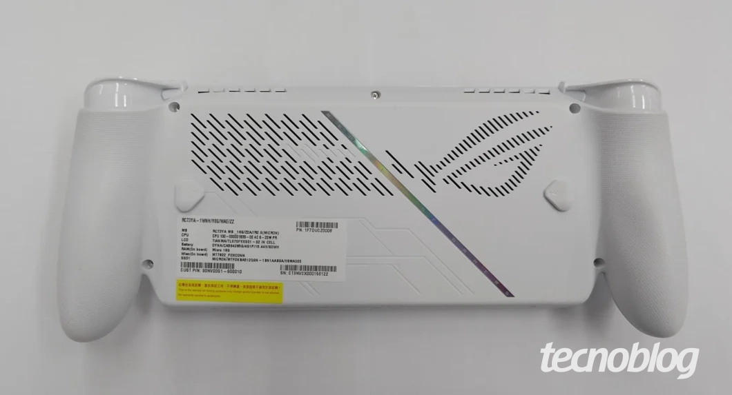 Foto em close-up da traseira do console portátil ROG Xbox Ally de cor branca. A parte central possui orifícios de ventilação dispostos em um padrão diagonal e um gráfico de arco-íris (holográfico) na lateral direita. Há pegadas ergonômicas texturizadas e um adesivo de informação da Anatel. O logo do "Tecnoblog" aparece no canto inferior direito.