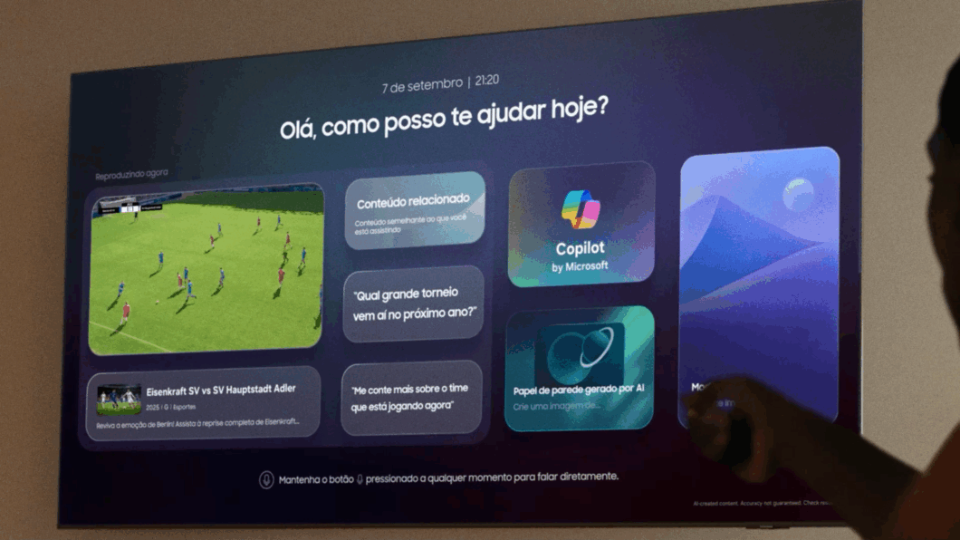 Tela de TV com interface de assistente virtual. No topo, aparecem a data e o hor&aacute;rio &ldquo;7 de setembro | 21:20&rdquo; e a frase &ldquo;Ol&aacute;, como posso te ajudar hoje?&rdquo;. &Agrave; esquerda, um jogo de futebol &eacute; exibido com o texto &ldquo;Reproduzindo agora&rdquo;. H&aacute; cart&otilde;es com sugest&otilde;es como &ldquo;Conte&uacute;do relacionado&rdquo;, &ldquo;Qual grande torneio vem a&iacute; no pr&oacute;ximo ano?&rdquo; e &ldquo;Me conte mais sobre o time que est&aacute; jogando agora&rdquo;. Tamb&eacute;m aparecem os &iacute;cones &ldquo;Copilot by Microsoft&rdquo; e &ldquo;Papel de parede gerado por AI&rdquo;. Uma pessoa aponta um controle remoto para a TV.