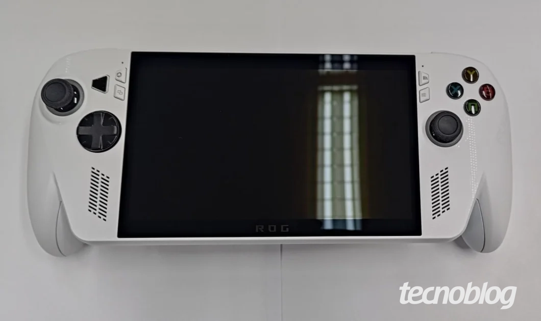 Foto superior do console de videogame portátil ROG Xbox Ally na cor branca. O dispositivo tem tela preta desligada que reflete uma janela. Nas laterais, há controles analógicos pretos, D-pad, botões de ação coloridos e fendas de ventilação. O logo "ROG" é visível na borda inferior da tela. O logo do "Tecnoblog" aparece no canto inferior direito.