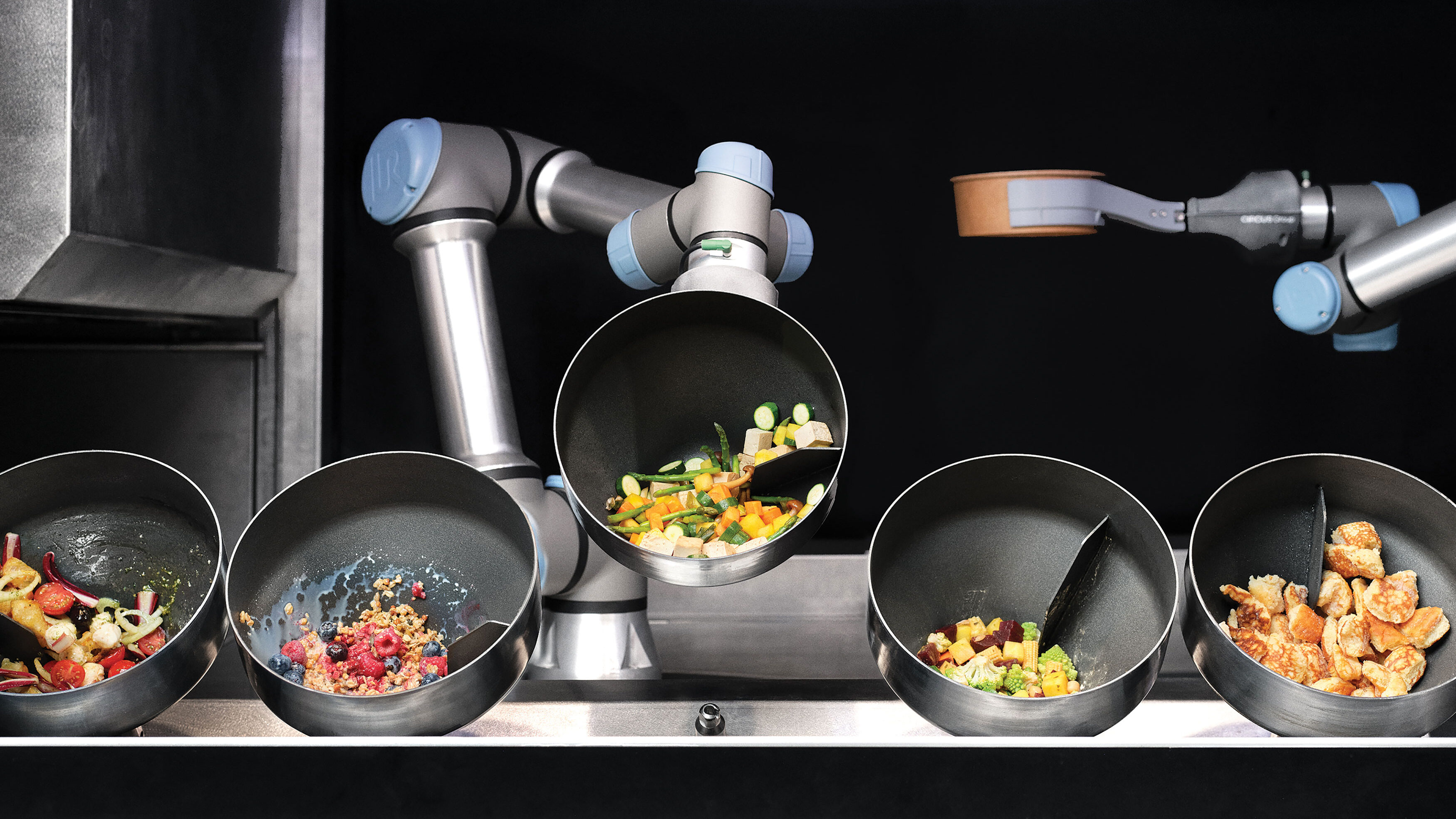 Braços robóticos metálicos cozinham alimentos em pequenas panelas inclinadas, alinhadas lado a lado. Cada panela contém ingredientes diferentes, como legumes picados, frutas, grãos e pedaços de carne. O ambiente lembra uma cozinha industrial automatizada, com foco na preparação de refeições sem intervenção humana.