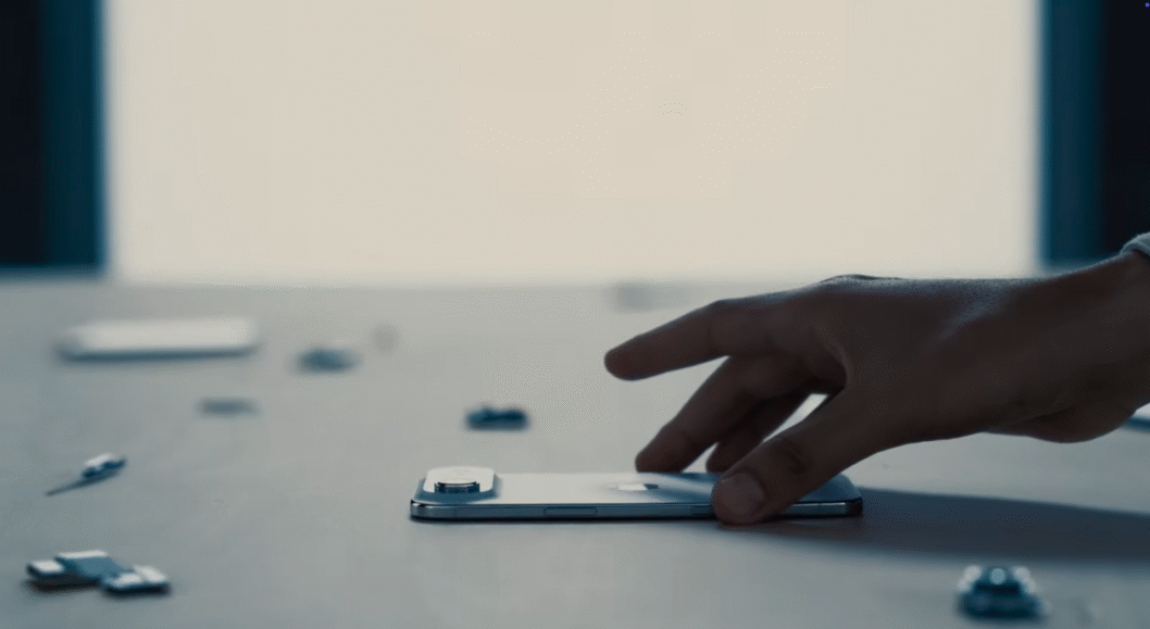 Foto em close-up de uma mão abaixando-se para pegar um iPhone Air prateado, que está deitado com a tela para baixo sobre uma superfície neutra.
