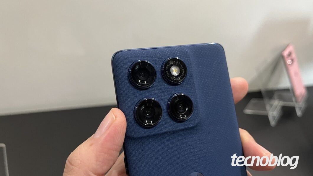 A imagem mostra a parte traseira do Motorola Edge 60, com foco na &aacute;rea das c&acirc;meras. O dispositivo possui um m&oacute;dulo de c&acirc;meras quadrado, com tr&ecirc;s lentes circulares e um flash LED. O aparelho &eacute; de cor azul escuro e est&aacute; sendo segurado com uma m&atilde;o. Ao fundo, &eacute; poss&iacute;vel ver um suporte transparente com outros aparelhos.