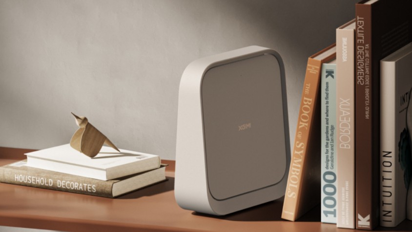 O projetor XGIMI Elfin Flip fechado, assemelhando-se a um livro ou pasta elegante de cor creme, posicionado verticalmente em uma prateleira ao lado de livros de arte e design. A imagem reforça a estética minimalista e o tamanho compacto do dispositivo.