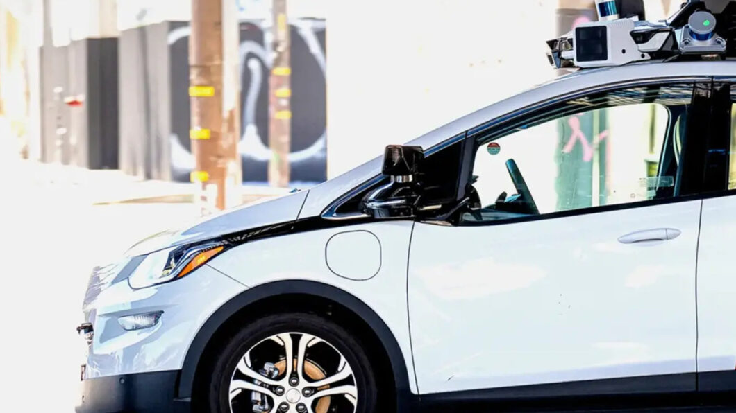 Carro el&eacute;trico branco estacionado em &aacute;rea de teste com equipamentos de sensor no teto, utilizado para opera&ccedil;&otilde;es de robotaxi com carregamento automatizado