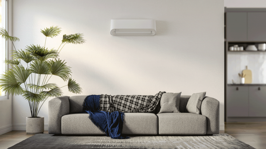 Ar-Condicionado Electrolux pendurado a uma parede branca. Embaixo um sof&aacute; e tapete na cor cinza e um vaso de planta ao lado.