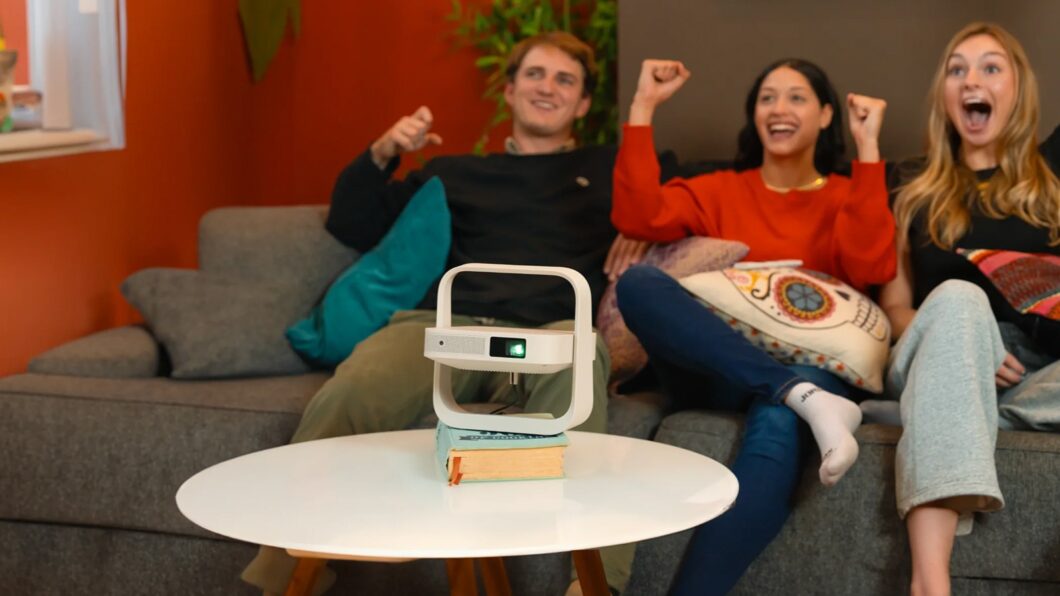 Cena de estilo de vida mostrando três amigos (um homem e duas mulheres) comemorando e vibrando em um sofá cinza. À frente deles, em uma mesa de centro branca e apoiado sobre um livro, o projetor XGIMI Elfin Flip branco está ligado, projetando luz para fora do quadro.