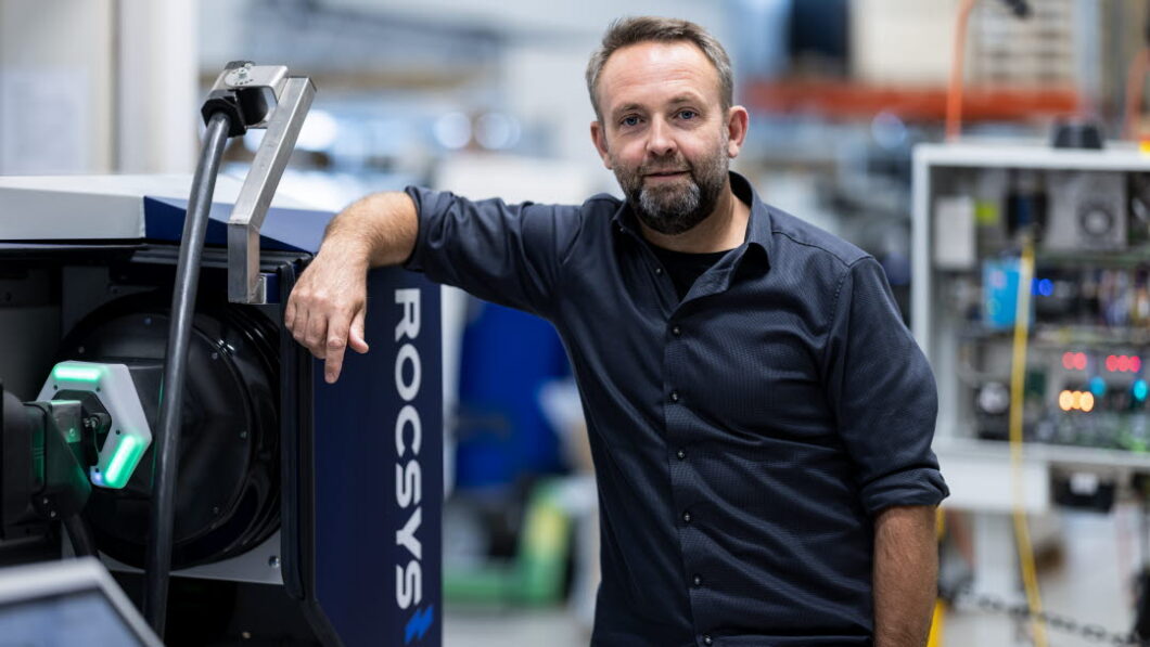 Imagem mostra um homem com camisa social azul escura em uma f&aacute;brica, com o bra&ccedil;o direito apoiado em um rob&ocirc; 