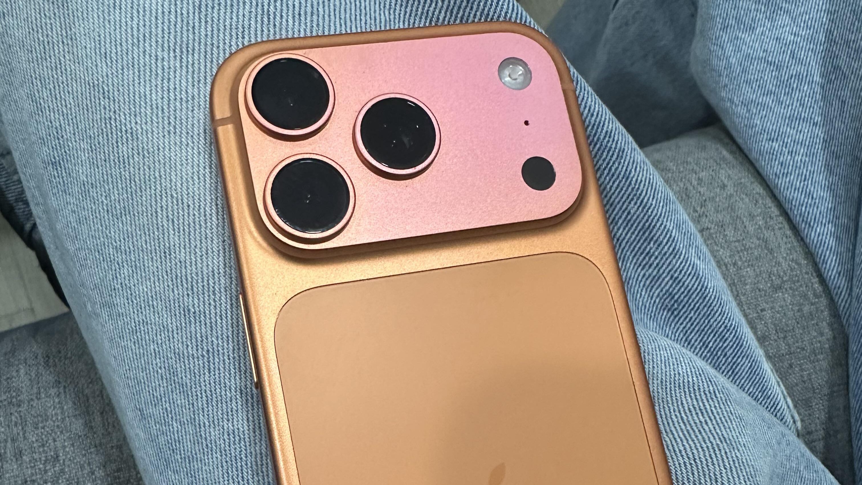 Visão da parte traseira de um mockup ou conceito do iPhone 17 Pro em dois tons, segurado sobre uma perna vestida com jeans azul claro. O design traseiro é dividido: o módulo de câmera (com três lentes escuras e sensores) é de um tom rosa metálico claro e o restante do painel traseiro é de um tom de cobre ou dourado pálido/alaranjado, com o logotipo da Apple ao centro.
