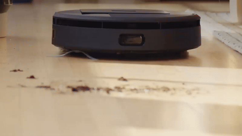 Gif animado mostra um robô aspirador Roomba, de cor preta, realizando limpeza em um piso de madeira