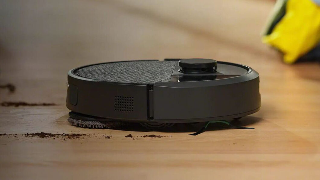 Imagem mostra um robô aspirador Roomba, de cor preta, realizando limpeza em um piso de madeira