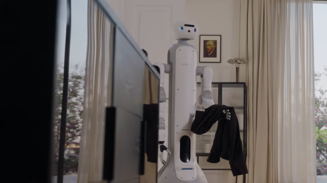 Rob&ocirc; humanoide branco e cinza, de estatura m&eacute;dia, em uma sala de estar iluminada. O rob&ocirc;, que tem o nome "onero" no peito, possui uma cabe&ccedil;a arredondada com grandes olhos azuis digitais. Ele segura uma pe&ccedil;a de roupa escura com sua m&atilde;o rob&oacute;tica direita. Ao fundo, v&ecirc;-se uma cortina clara, um quadro na parede e um arm&aacute;rio com objetos decorativos. A composi&ccedil;&atilde;o &eacute; um plano m&eacute;dio que destaca a intera&ccedil;&atilde;o do rob&ocirc; com o ambiente dom&eacute;stico.