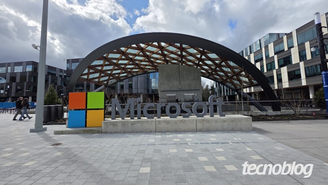 Imagem da entrada do campus da Microsoft em Redmond, nos Estados Unidos. Em destaque, o letreiro com o nome "Microsoft" ao lado do logo colorido da empresa. Ao fundo, h&aacute; um edif&iacute;cio moderno com uma cobertura de madeira e vidro em arco. O c&eacute;u est&aacute; parcialmente nublado e algumas pessoas caminham pela cal&ccedil;ada.